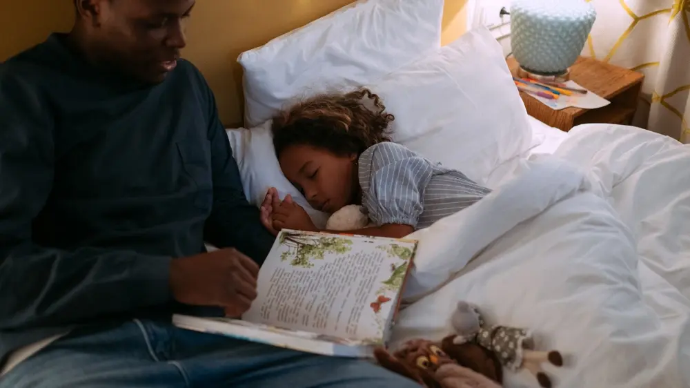 Parent reading aloud beside a child settling into bed with a stuffed animal and a lamp on the nightstand